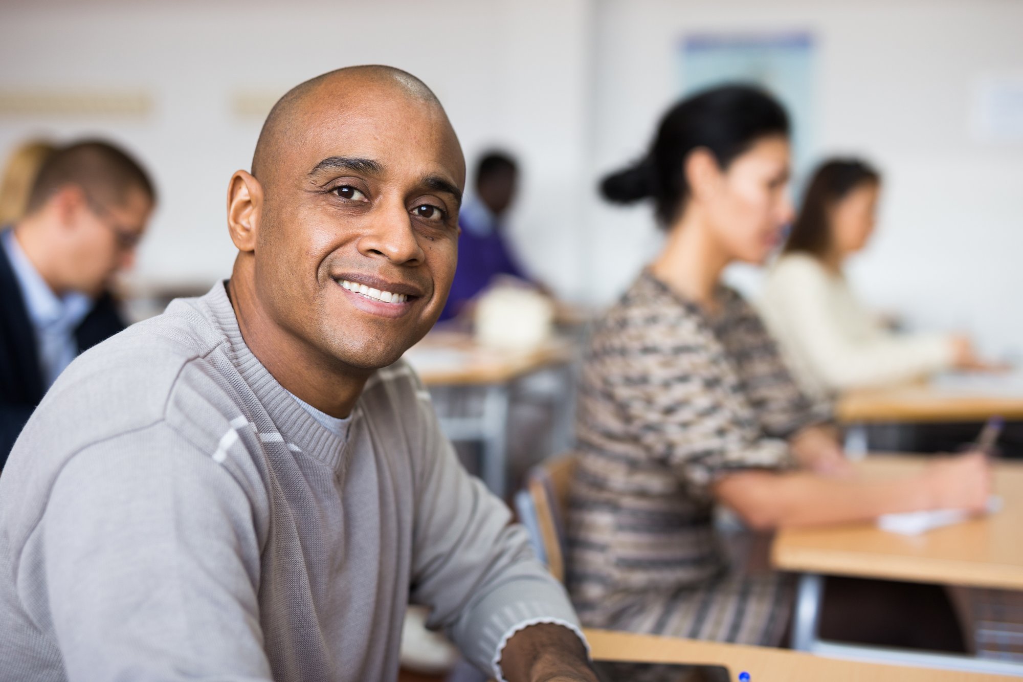 A bald man smiling at the camera in a classroom with other students focused on writing behind him. A bald man smiling at the camera in a classroom with other students focused on writing behind him.