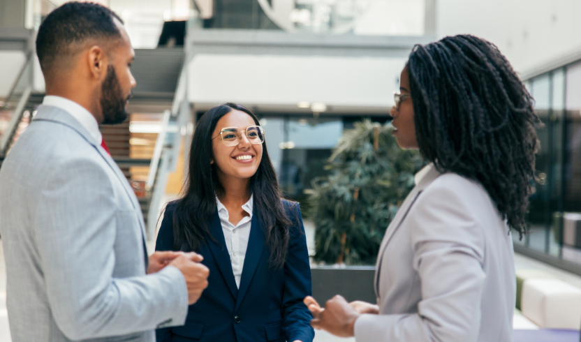 Three business professionals in formal attire engage in a conversation in a modern office lobby. Two women and one man exchanging ideas and smiling.