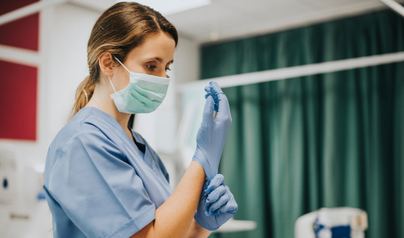 Female healthcare worker in scrubs and a mask putting on blue gloves in a hospital room.