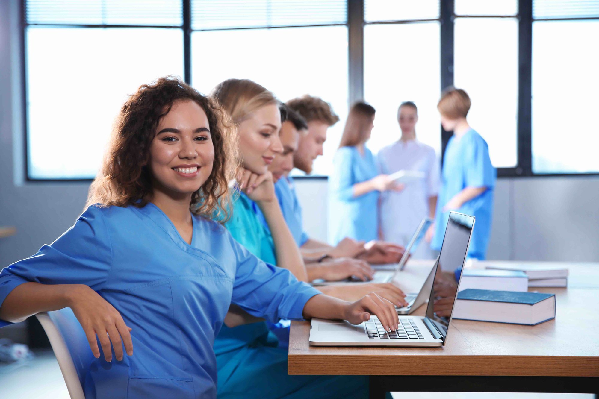 Medical students in scrubs using laptops in a classroom with large windows, one female student smiling at the camera.