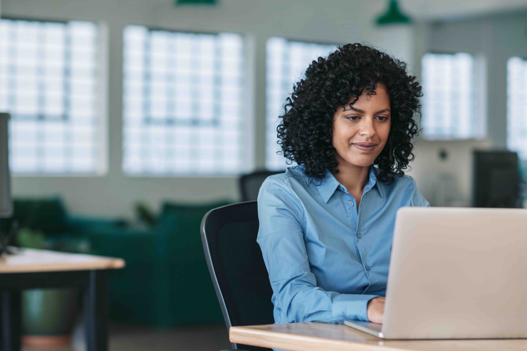 A woman with curly hair working on a laptop in a bright office space.