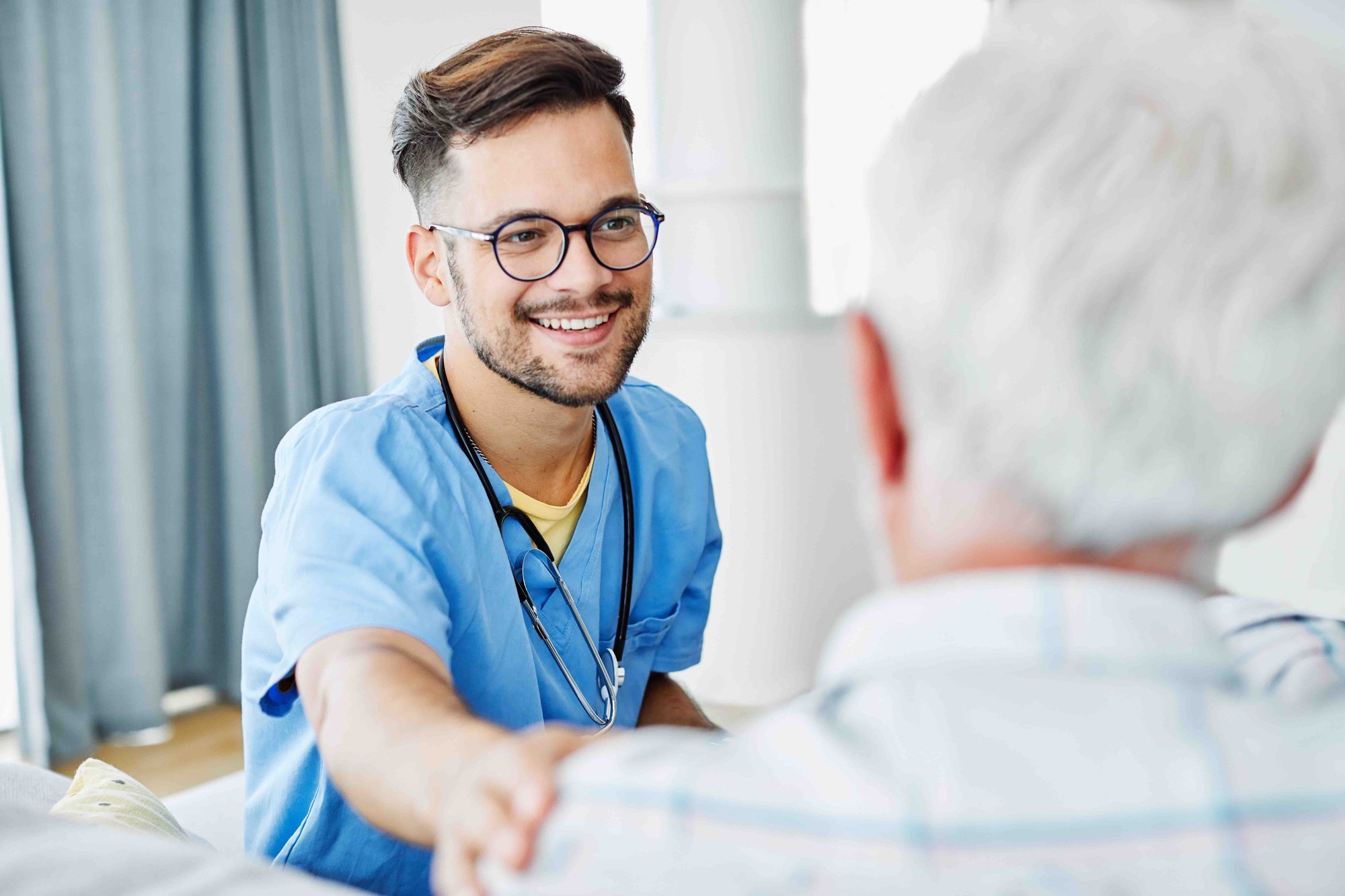 A smiling male nurse in blue scrubs shakes hands with an elderly patient, creating a warm and caring atmosphere in a bright hospital room.