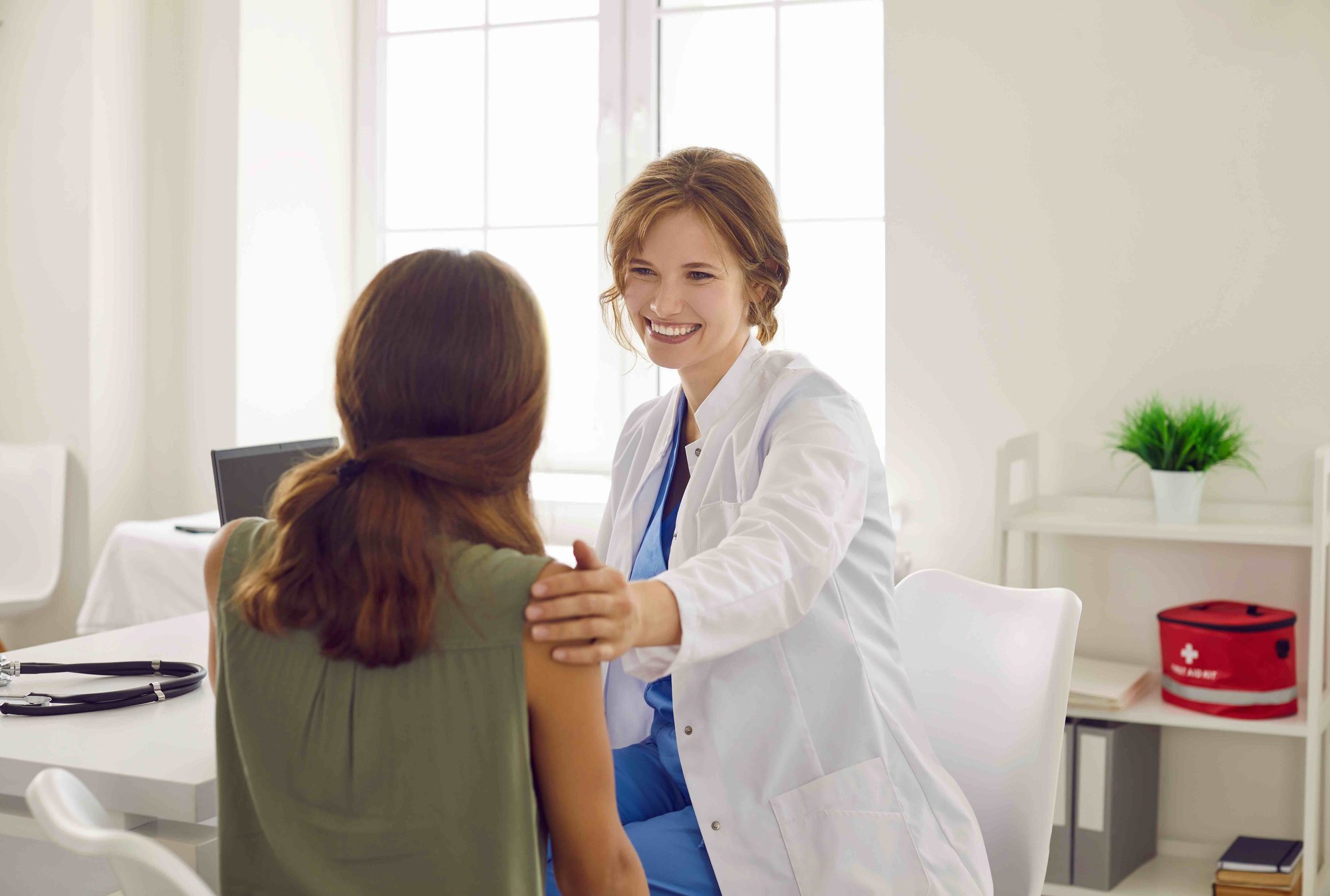 Female doctor with a stethoscope around her neck smiling at a female patient in a medical office, both sitting.