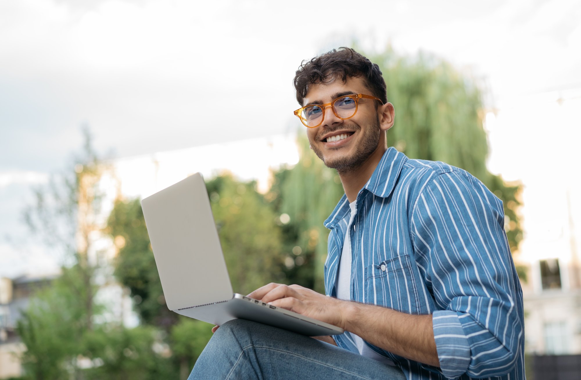 A young man wearing glasses and a striped shirt smiles while using a laptop outdoors.