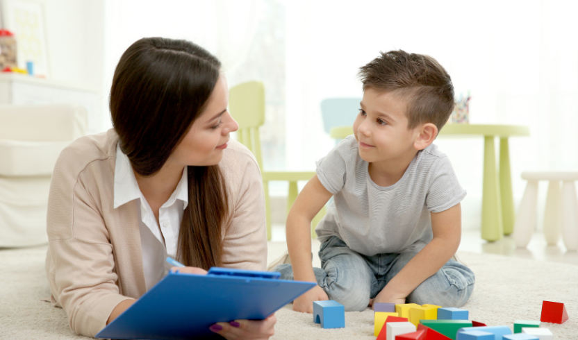 A woman with a clipboard engages with a child inside a brightly lit room. The child sits on the floor, surrounded by various colorful toy blocks.