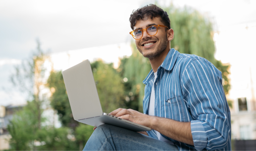 A man with glasses and a striped shirt smiles while working on a laptop outdoors on a sunny day. Trees and buildings are visible in the background.