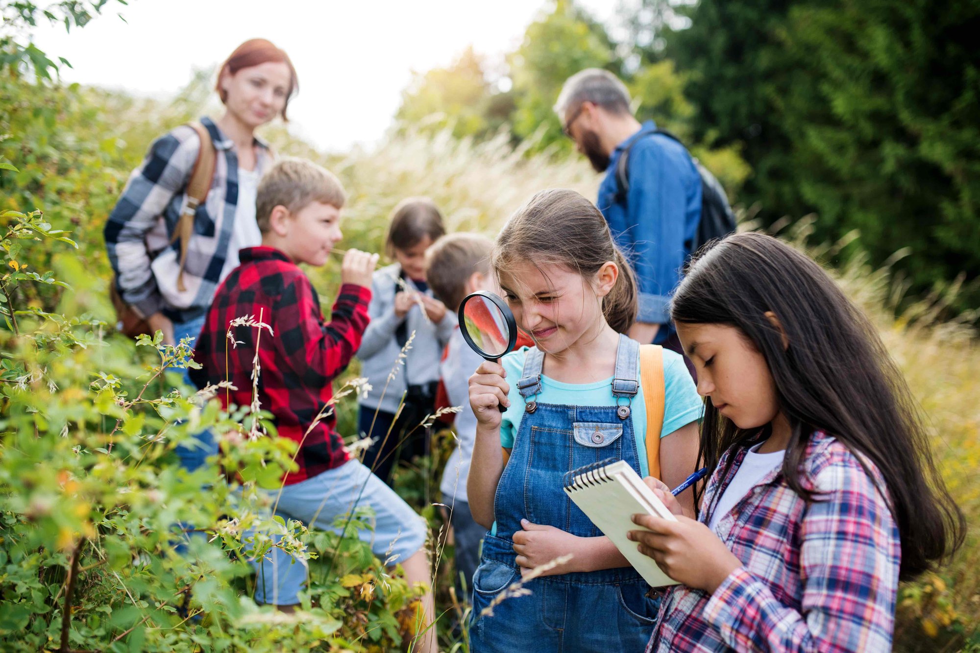 Group of children and adults exploring the outdoors. One child uses a magnifying glass to examine a plant, another takes notes in a notebook.