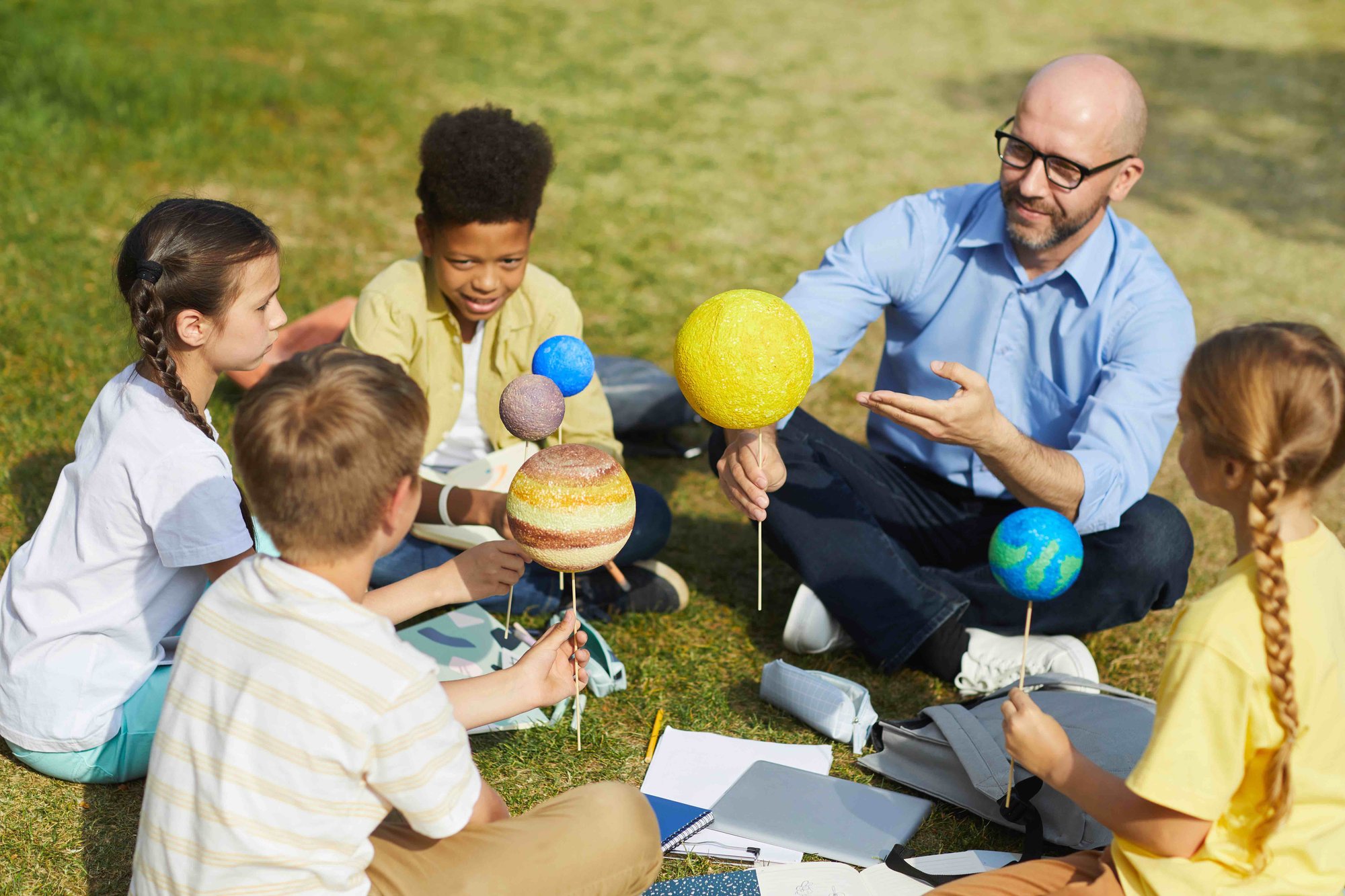 A teacher and four children sit on the grass while holding solar system model planets on sticks during an outdoor science lesson.