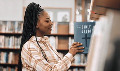 A woman with long braids reaches for a Bible Studies book in a library. She wears a plaid shirt and smiles as she looks at the book.