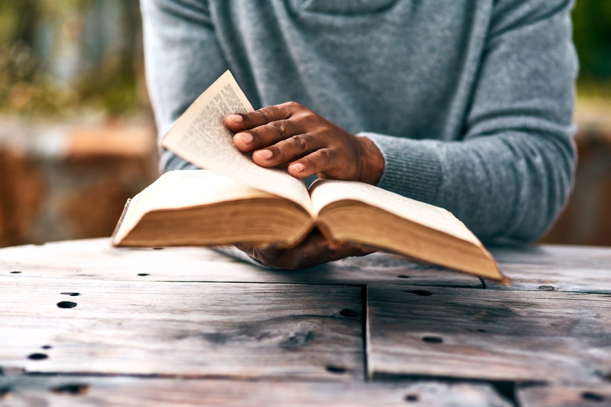 Person sitting at a wooden table reading an open book, focusing on their hands resting on the pages.