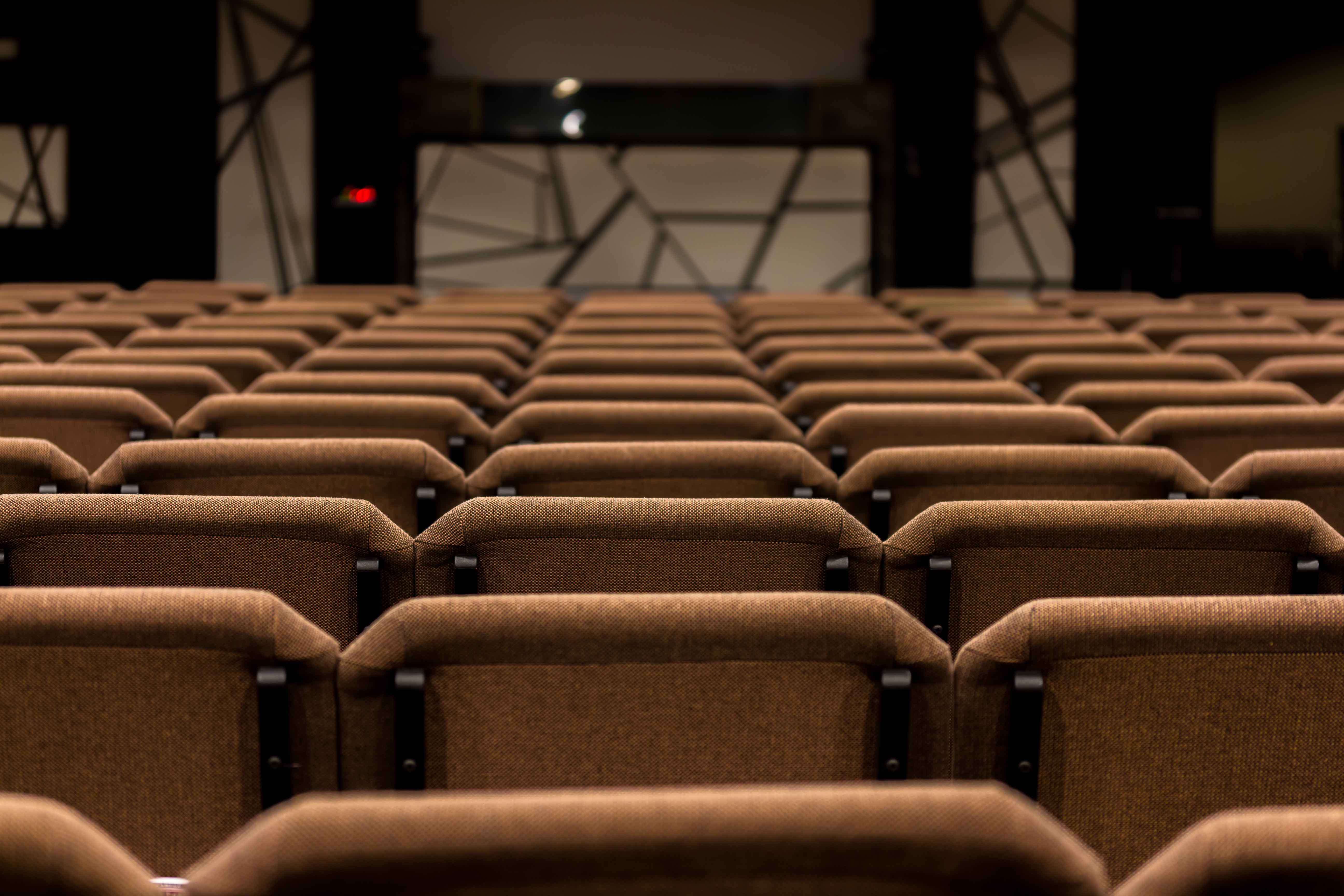 Rows of empty brown seats in an auditorium with a dark stage in the background.