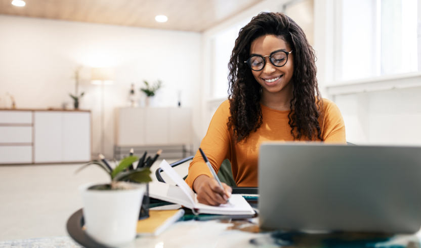 A woman with glasses and curly hair, wearing an orange top, is sitting at a table with a laptop and writing in a notebook.