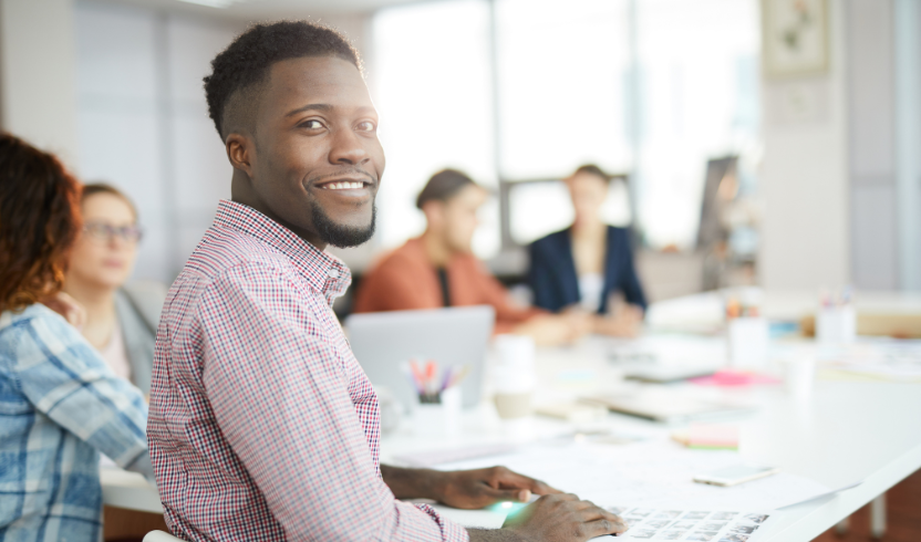 A man sits at a desk in an office environment, smiling at the camera. Other people are working in the background.