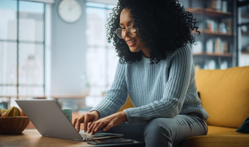 A woman with curly hair and glasses is sitting on a yellow sofa, working on a laptop with a bookshelf and windows in the background.